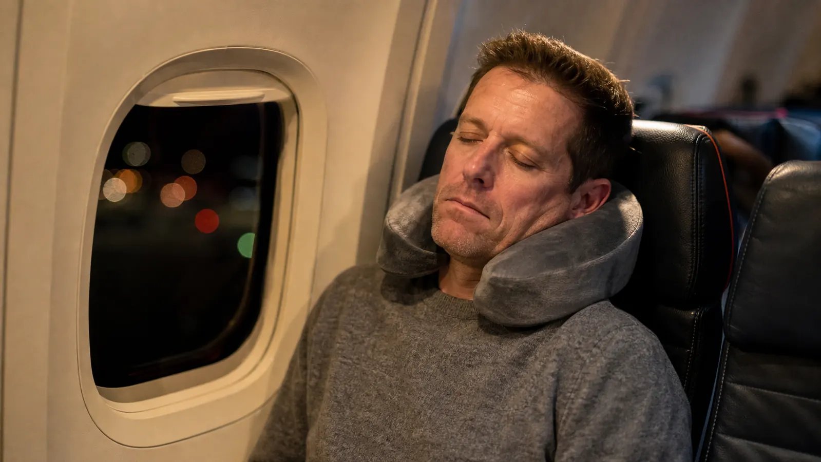 Traveler asleep in an airplane window seat wearing a gray neck pillow, soft cabin light