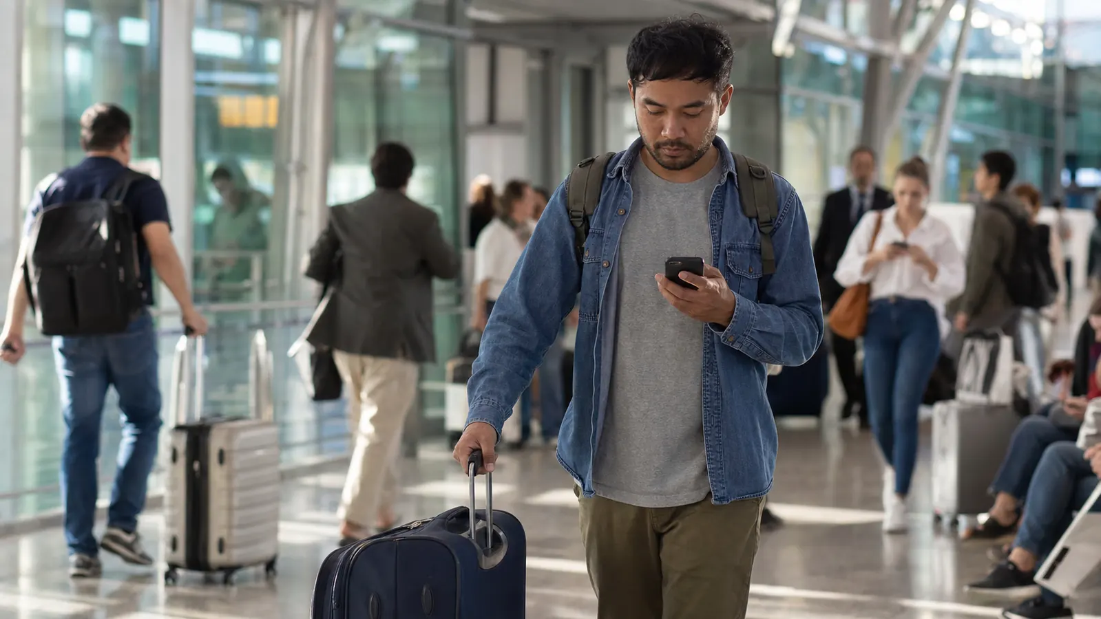 Traveler walking through an airport terminal checking a phone at forty percent battery while pulling a rolling carry-on
