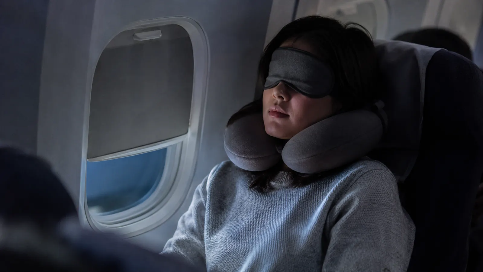 Traveler asleep in an economy window seat wearing a memory foam neck pillow during a dim red-eye flight