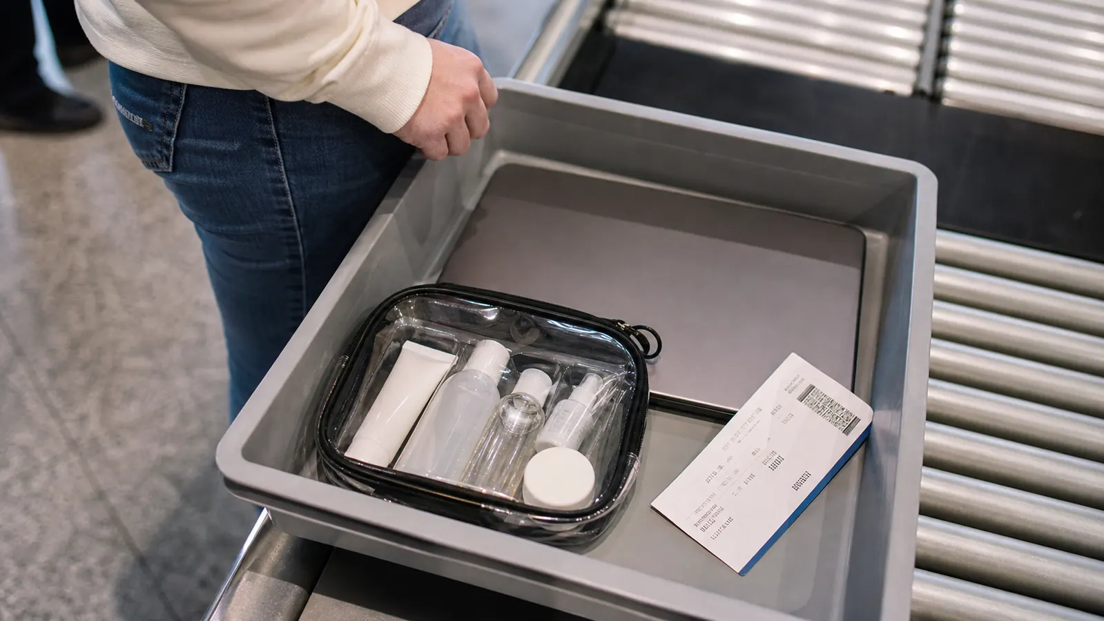 Clear quart-size toiletry bag with travel bottles sitting open on an airport security tray next to a boarding pass
