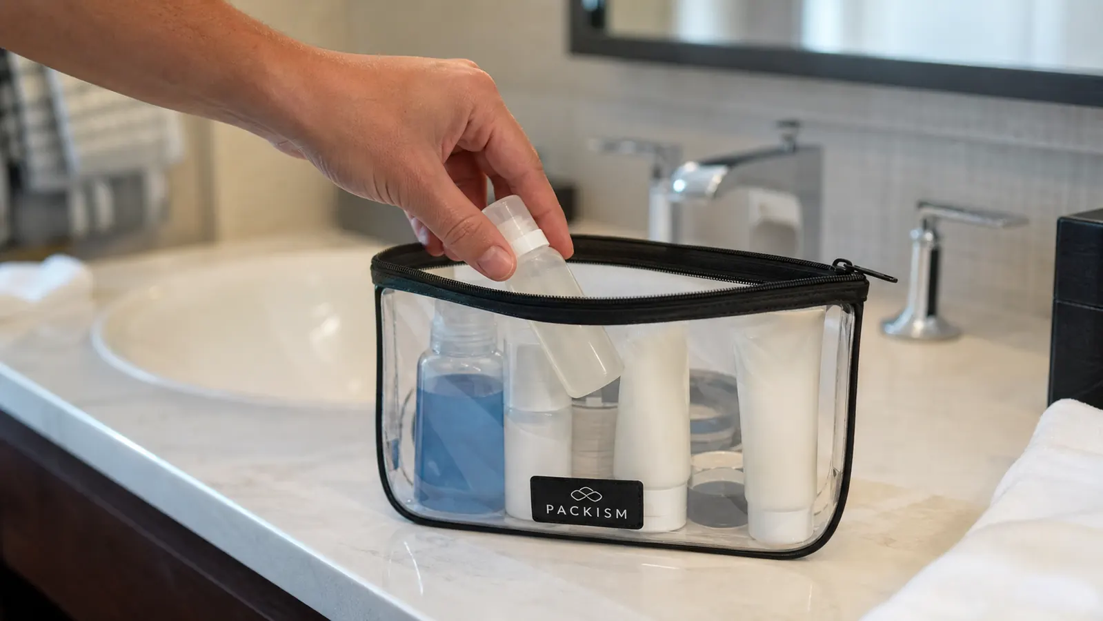 Hand placing travel-size bottles into a clear zippered toiletry bag on a bathroom counter
